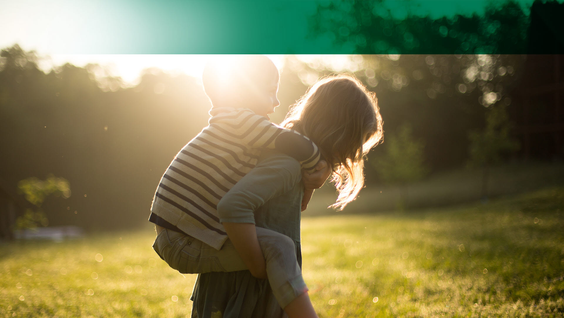 Young adult woman carrying young, smiling boy on her back while outside with sun shining in the background. 