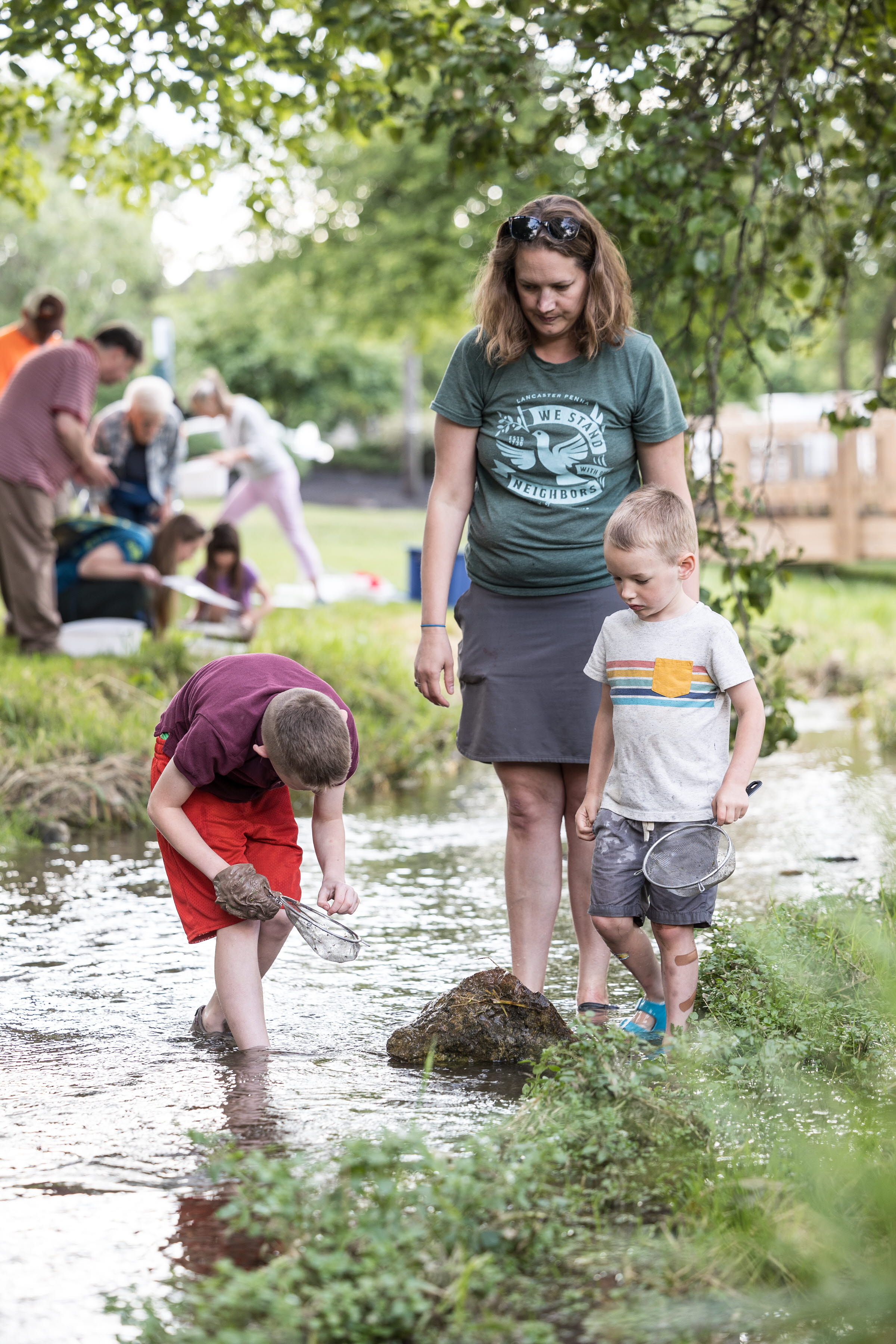 Adult white woman watching her two young children explore a stream 