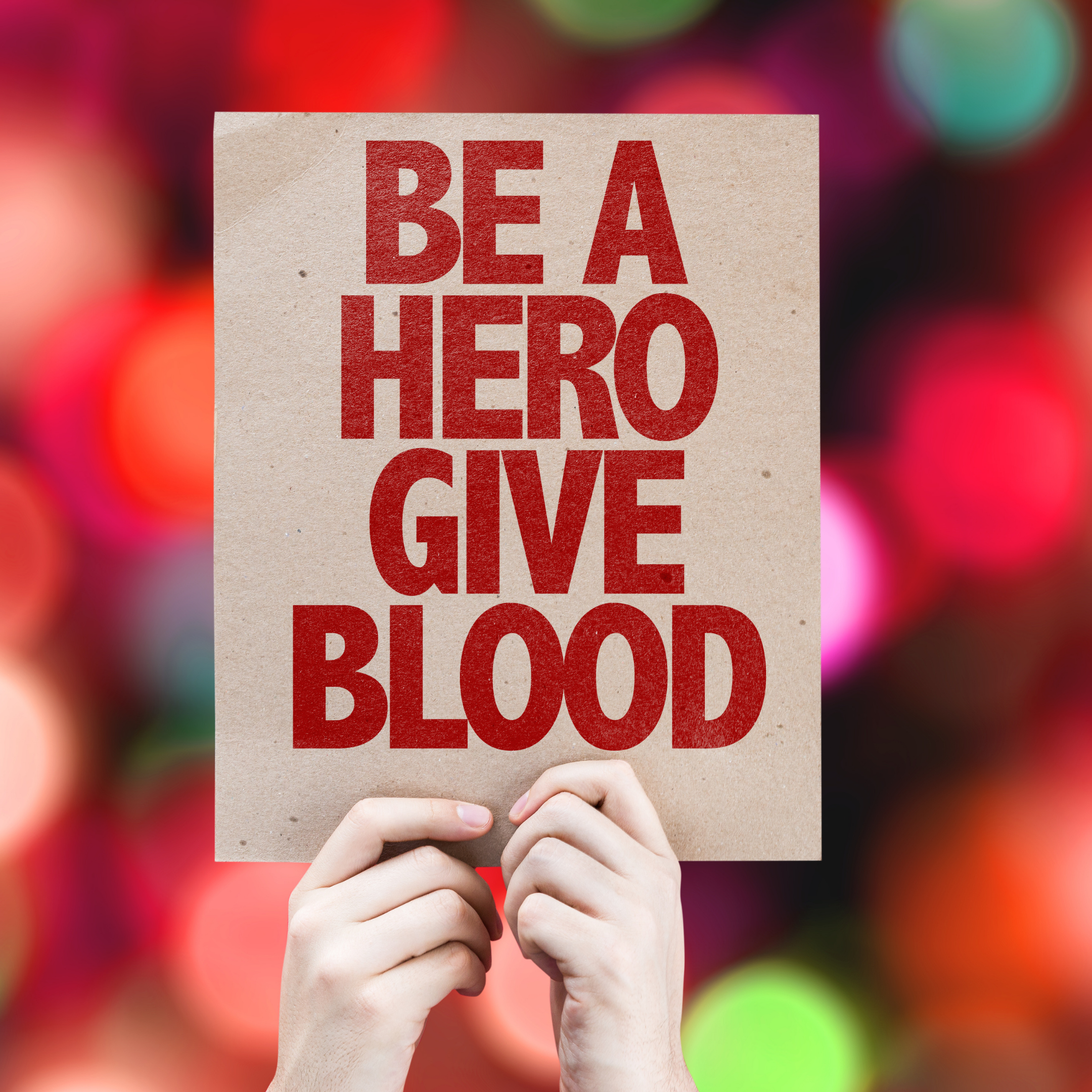 Hands holding up a sign that reads "Be a Hero Give Blood." 